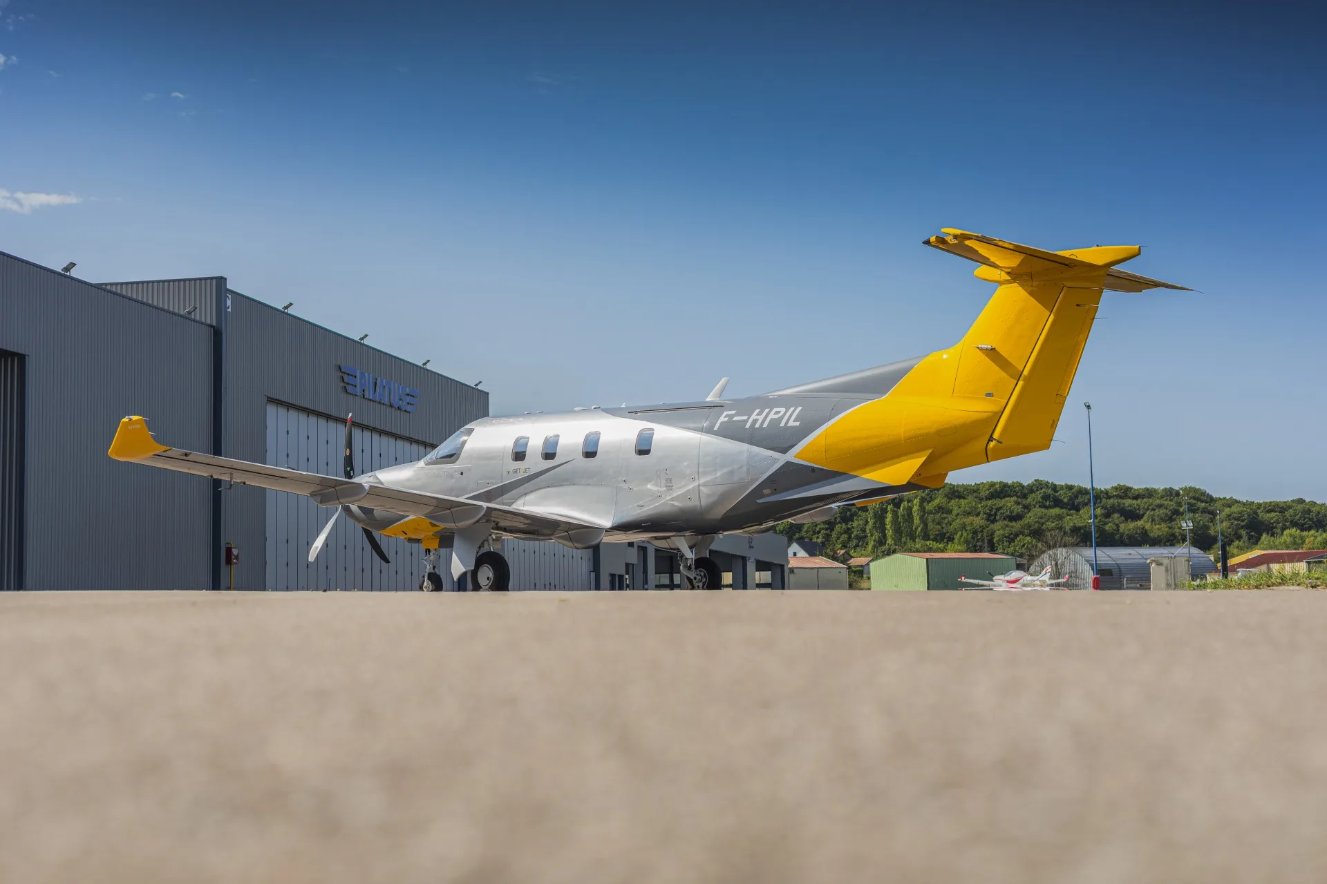 A silver and yellow Pilatus PC-12 aircraft (F-HPIL) parked on the tarmac in front of a hangar.