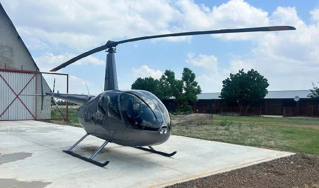 A front-left view of a dark gray Robinson R44 helicopter parked on a concrete pad next to a hangar.