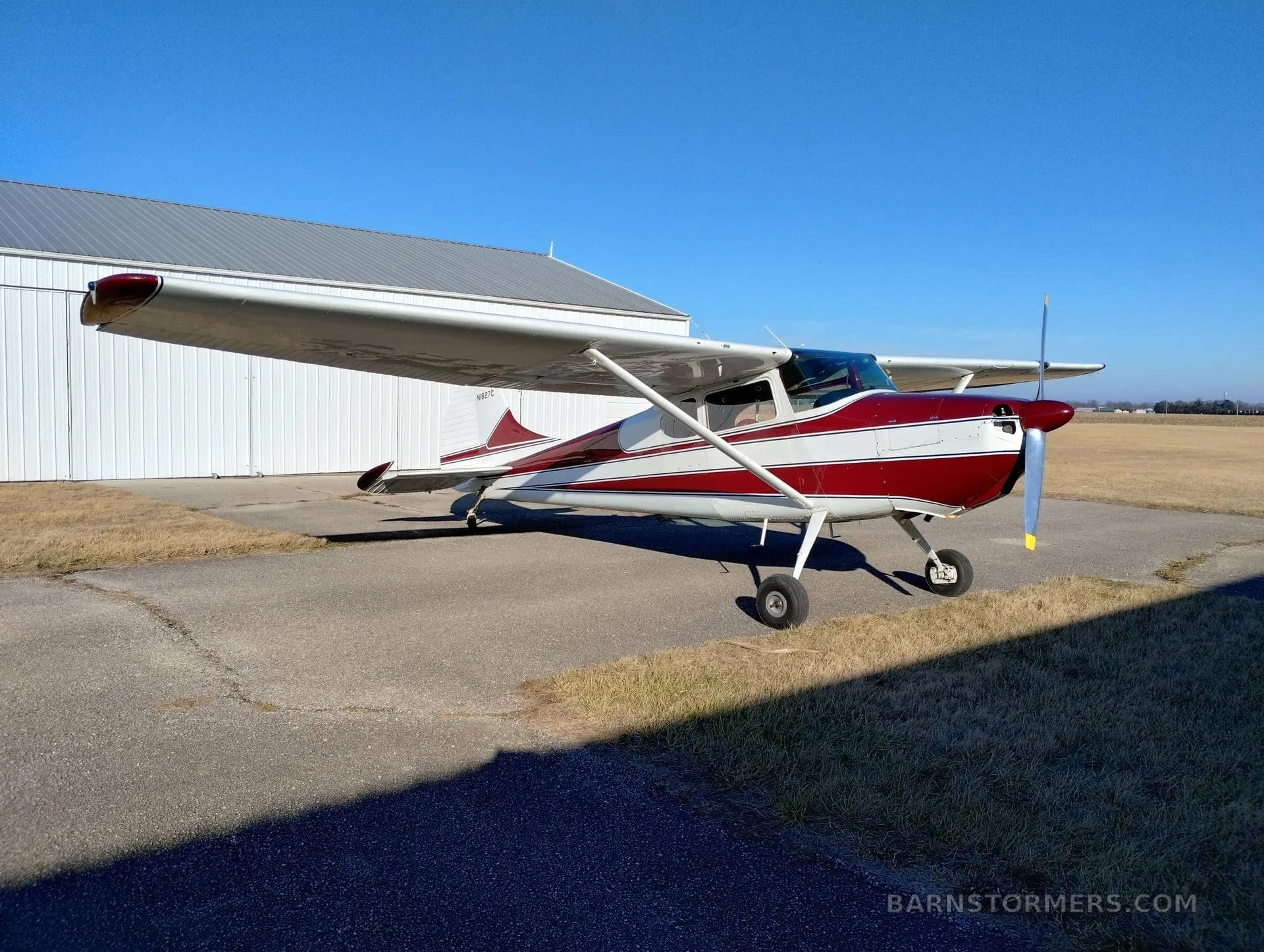 A red and white single-engine propeller aircraft is parked on an asphalt pad next to a white hangar under a clear blue sky.