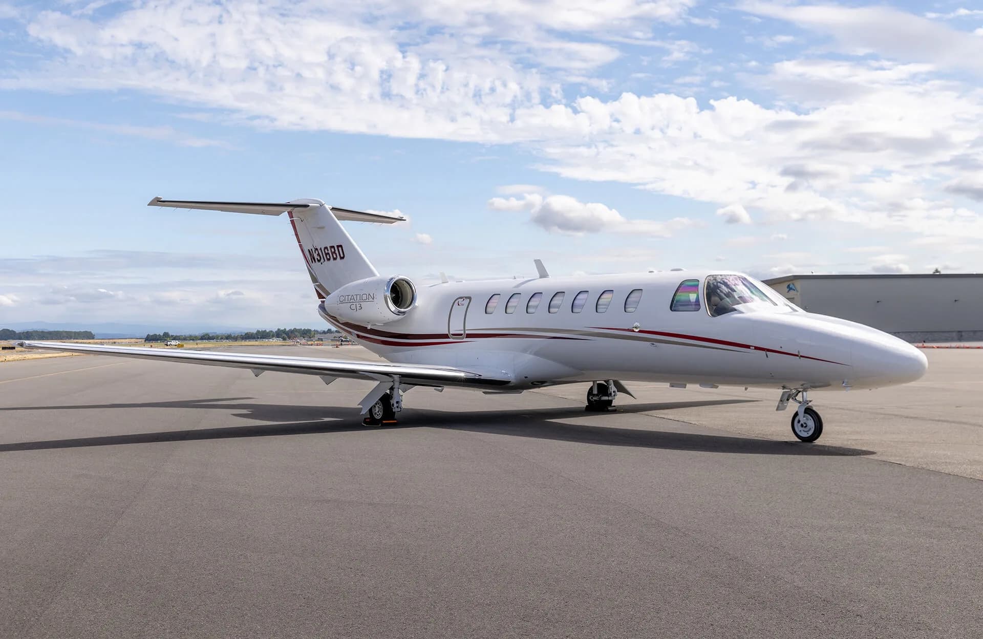 A white Cessna Citation CJ3 business jet with red and grey stripes is parked on an airport tarmac under a partly cloudy sky.