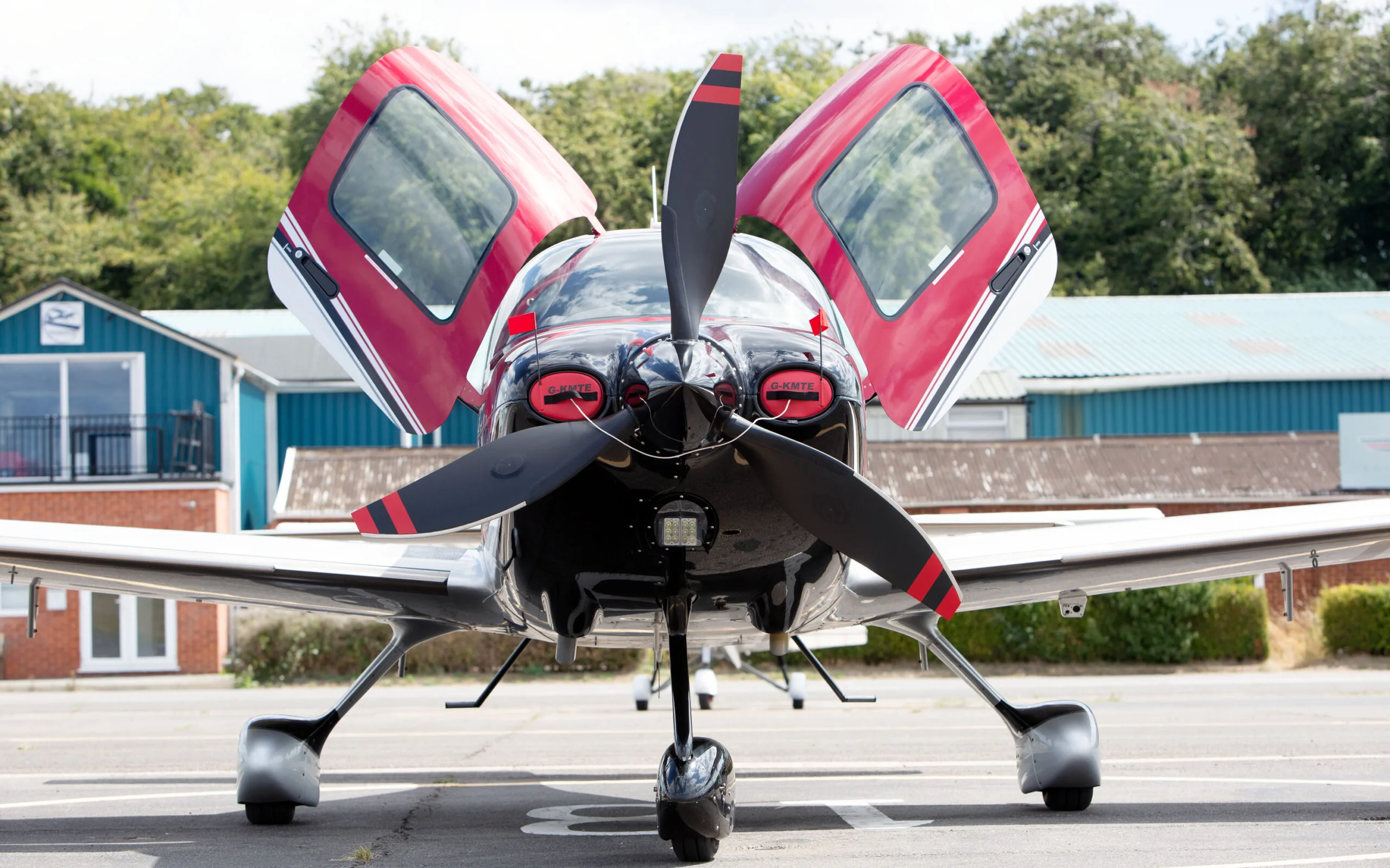A head-on view of a red, black, and silver Cirrus SR22 aircraft with its gull-wing doors open, parked on an airfield.