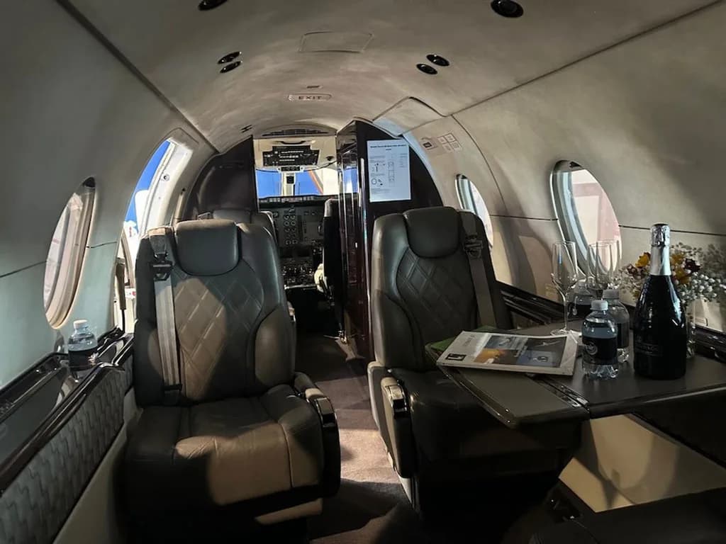 The interior cabin of a private jet with grey leather seats, a table with drinks, and a view towards the cockpit.