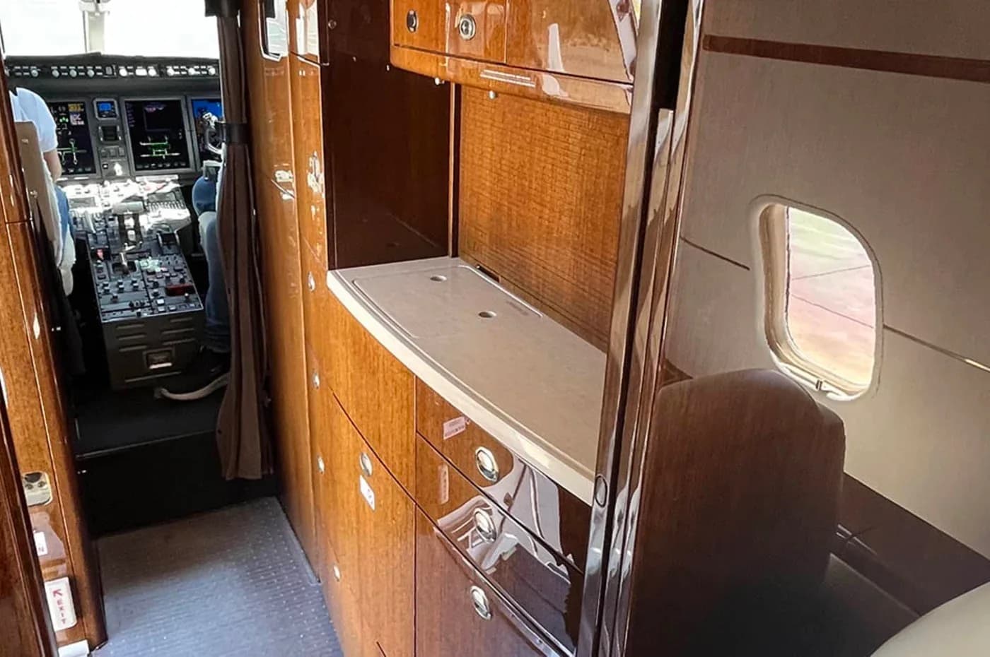 The galley area of a business jet, featuring polished wooden cabinetry, a countertop, and a glimpse of the cockpit in the background.