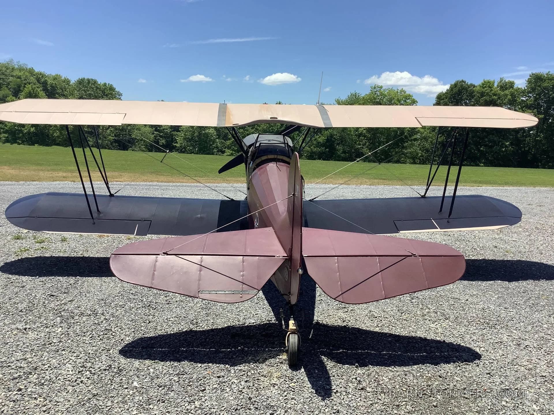 Rear view of a maroon and black biplane parked on a gravel surface under a blue sky.