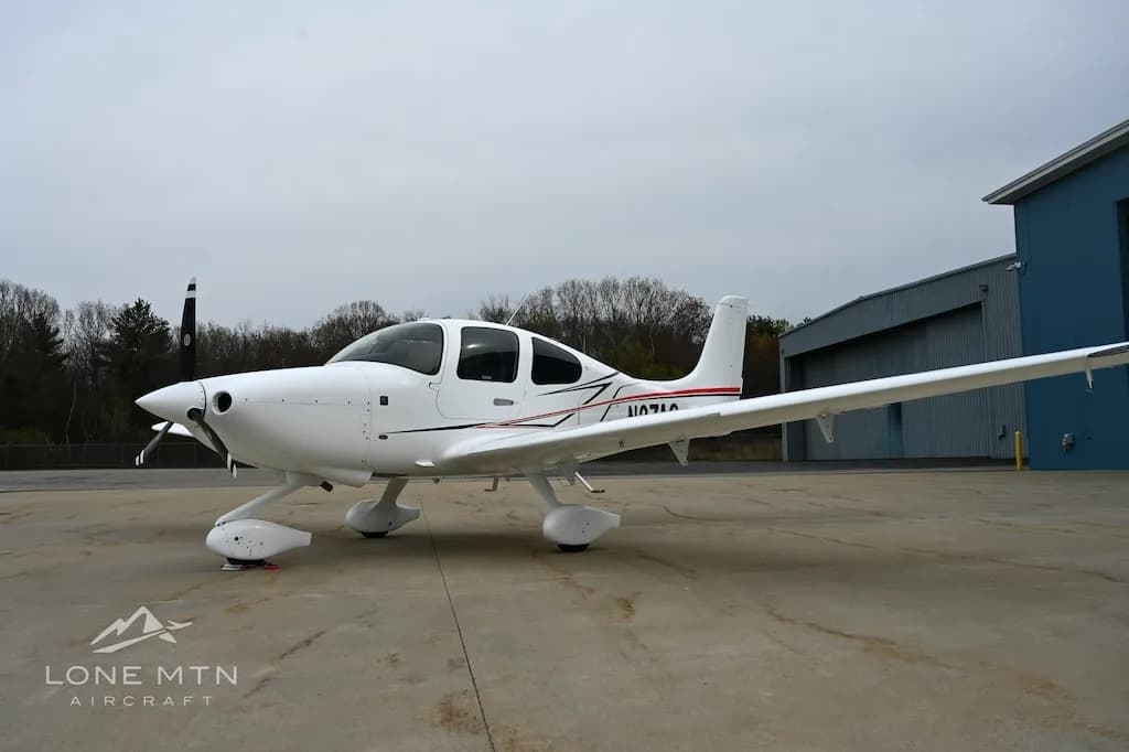 White Cirrus SR22 aircraft with red and black stripes, tail number N1071A, parked on an apron with trees in the background.