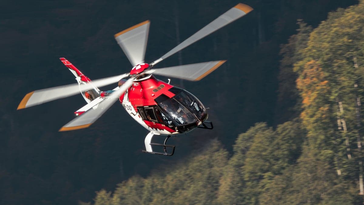A red and white Airbus H135 helicopter in flight over a forest.
