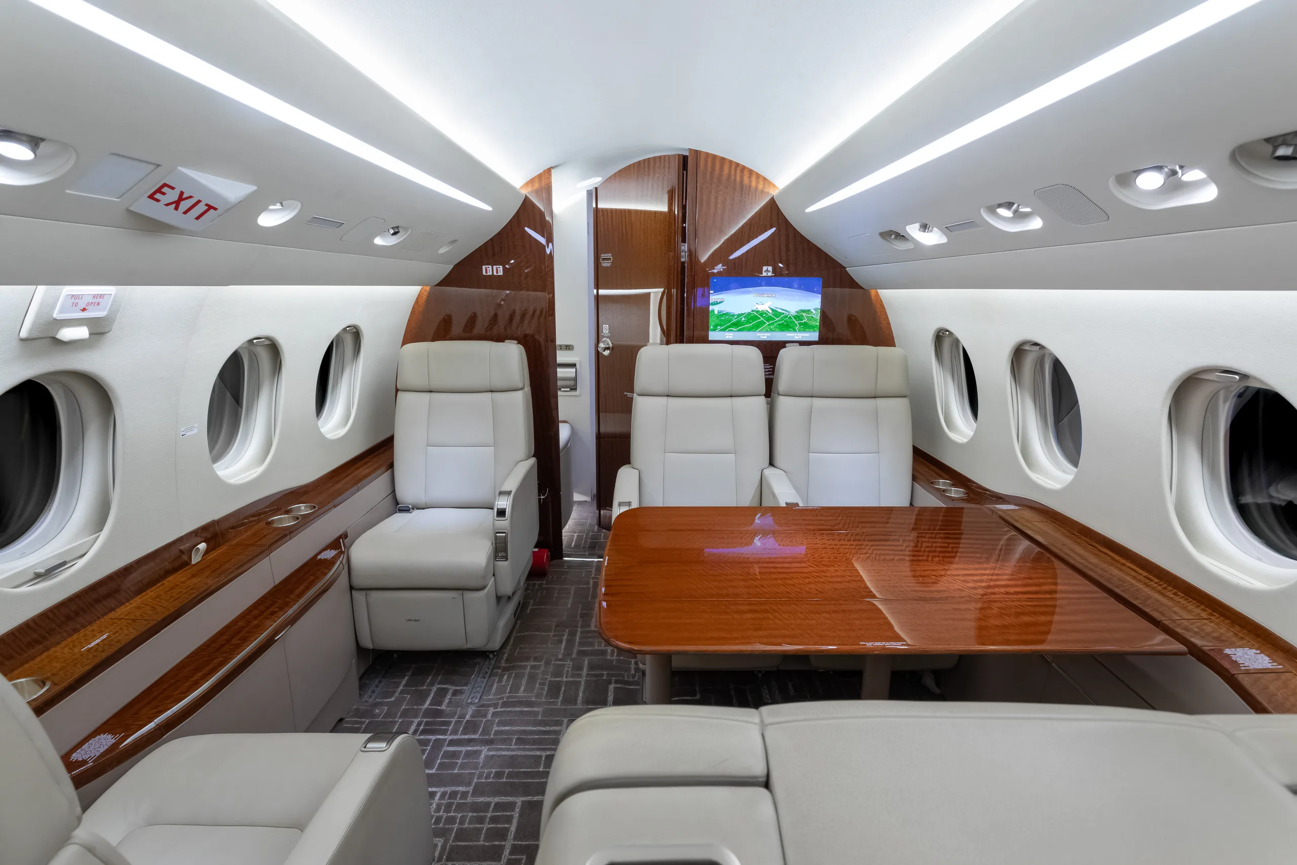 View of a luxurious aircraft cabin showing light beige leather seats, a large polished wooden table, and an "EXIT" sign above a window.