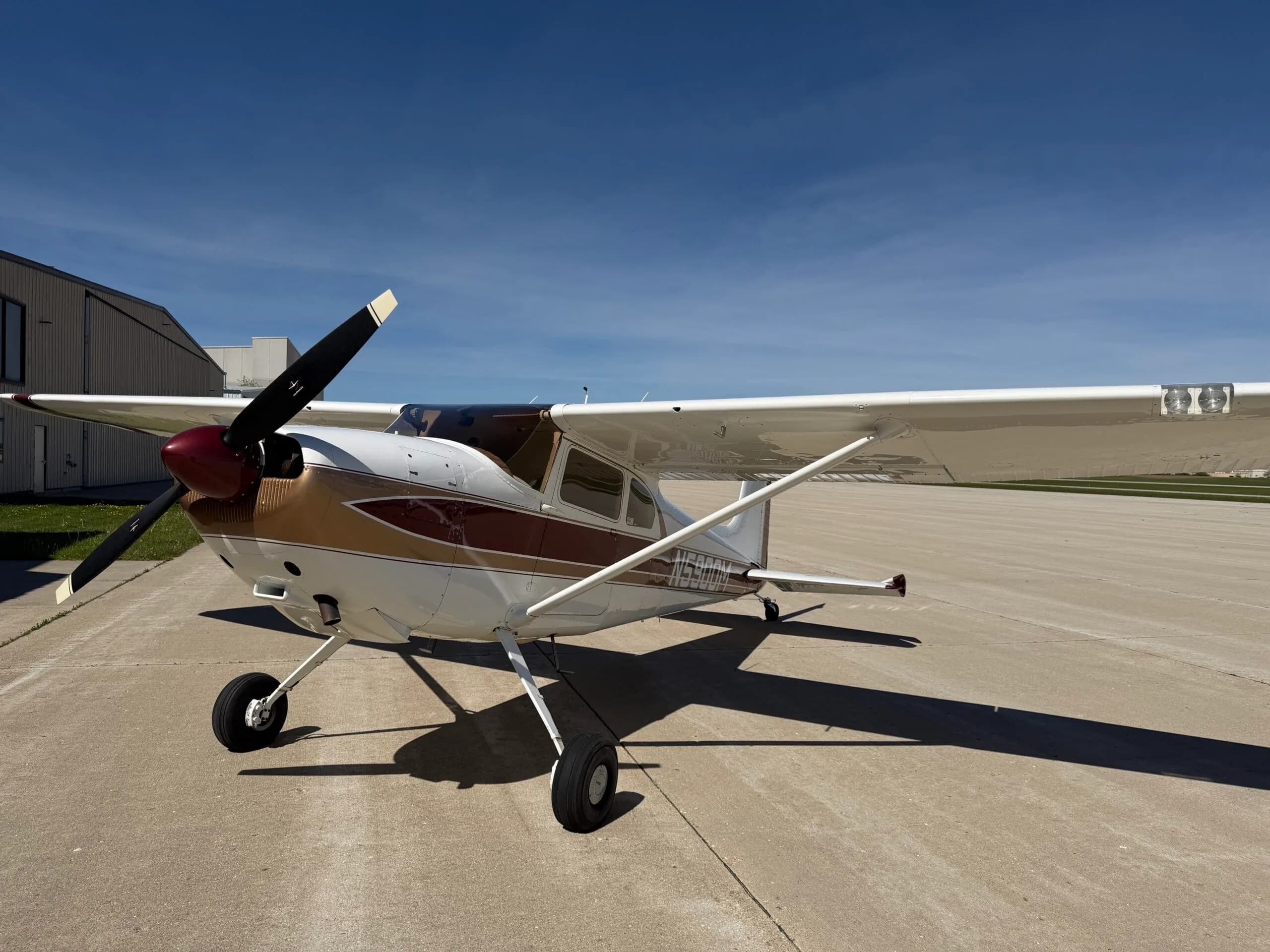 Front-left view of a white, gold, and maroon Cessna 172 parked on an airfield next to a hangar.