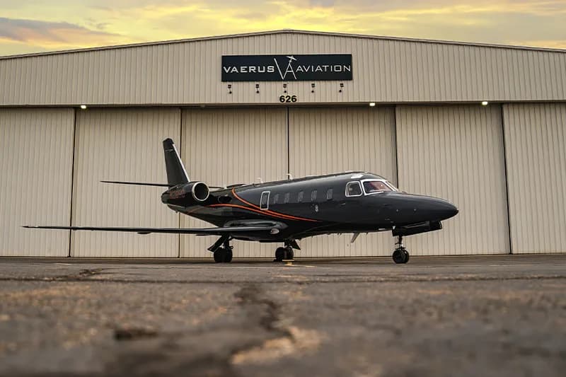 A dark gray jet with orange stripes parked in front of a hangar with the Vaerus Aviation logo.
