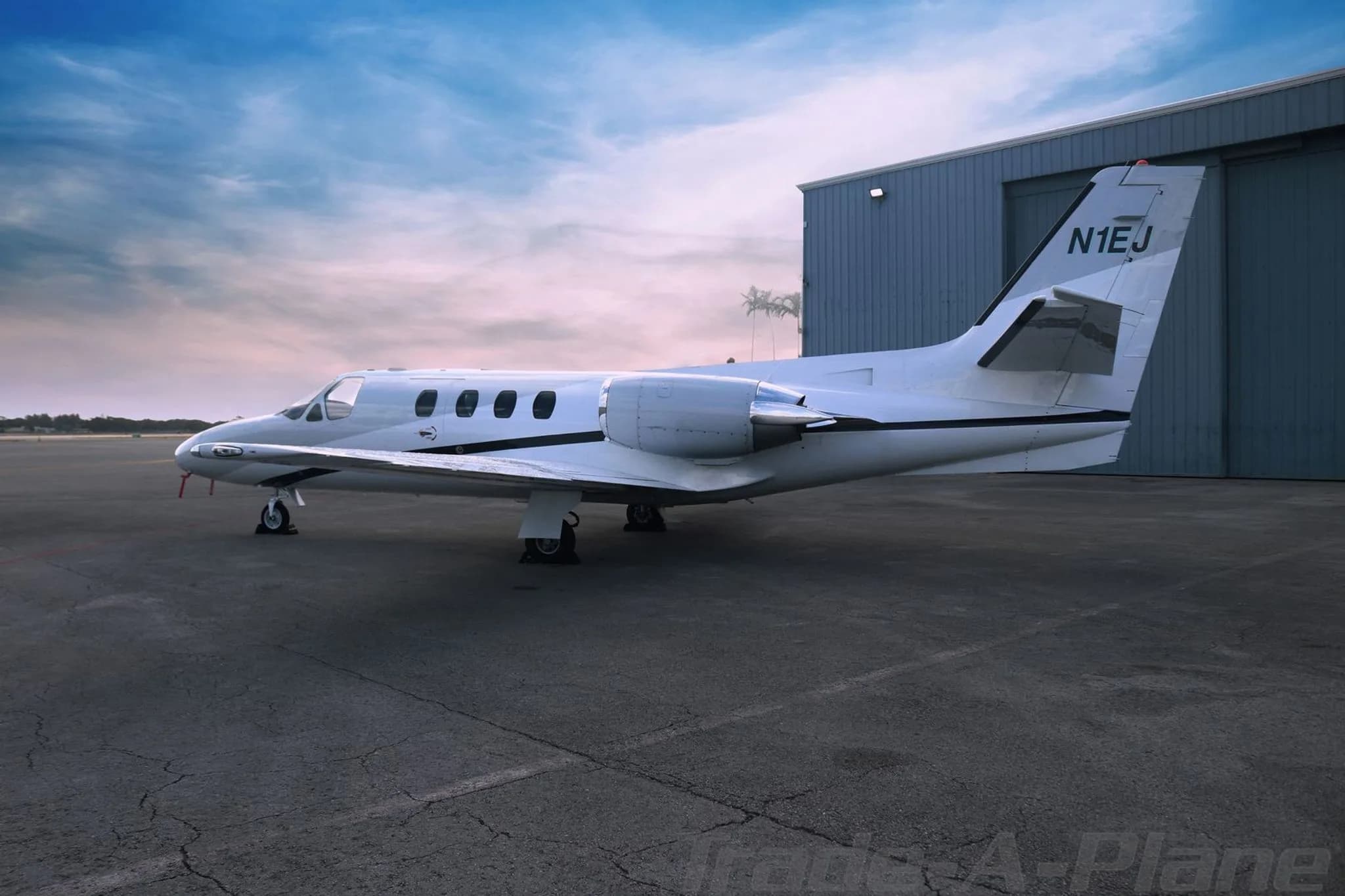 A side view of the white Cessna CitationJet N1EJ parked on an tarmac next to a blue hangar.