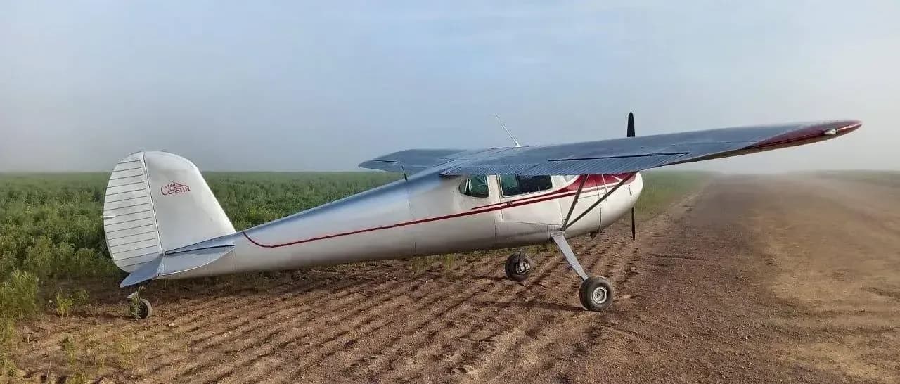A silver and red Cessna 170 aircraft is parked on a dirt field.