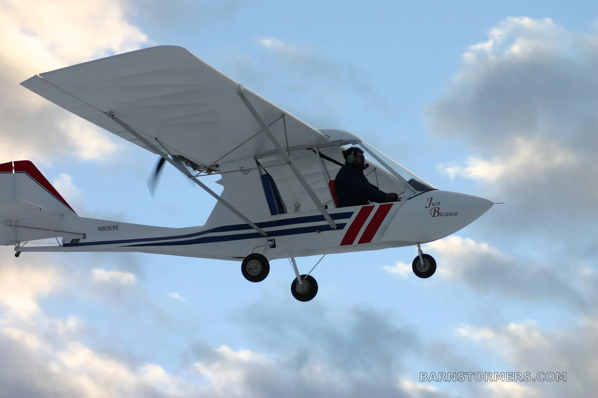 A white ultralight aircraft with blue and red stripes and "Just Because" on the side, flying against a cloudy sky.