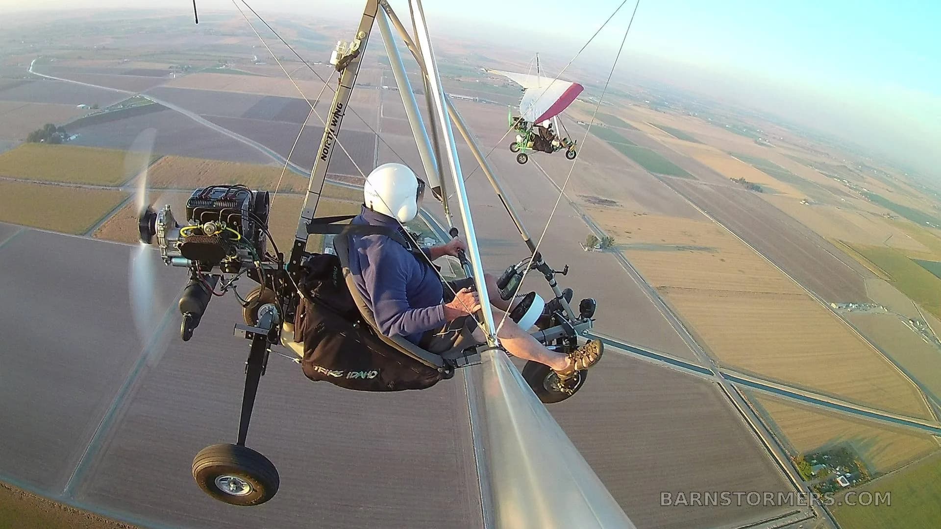 A pilot in a blue shirt flies a black and white ultralight aircraft over fields, with another ultralight flying in the distance.