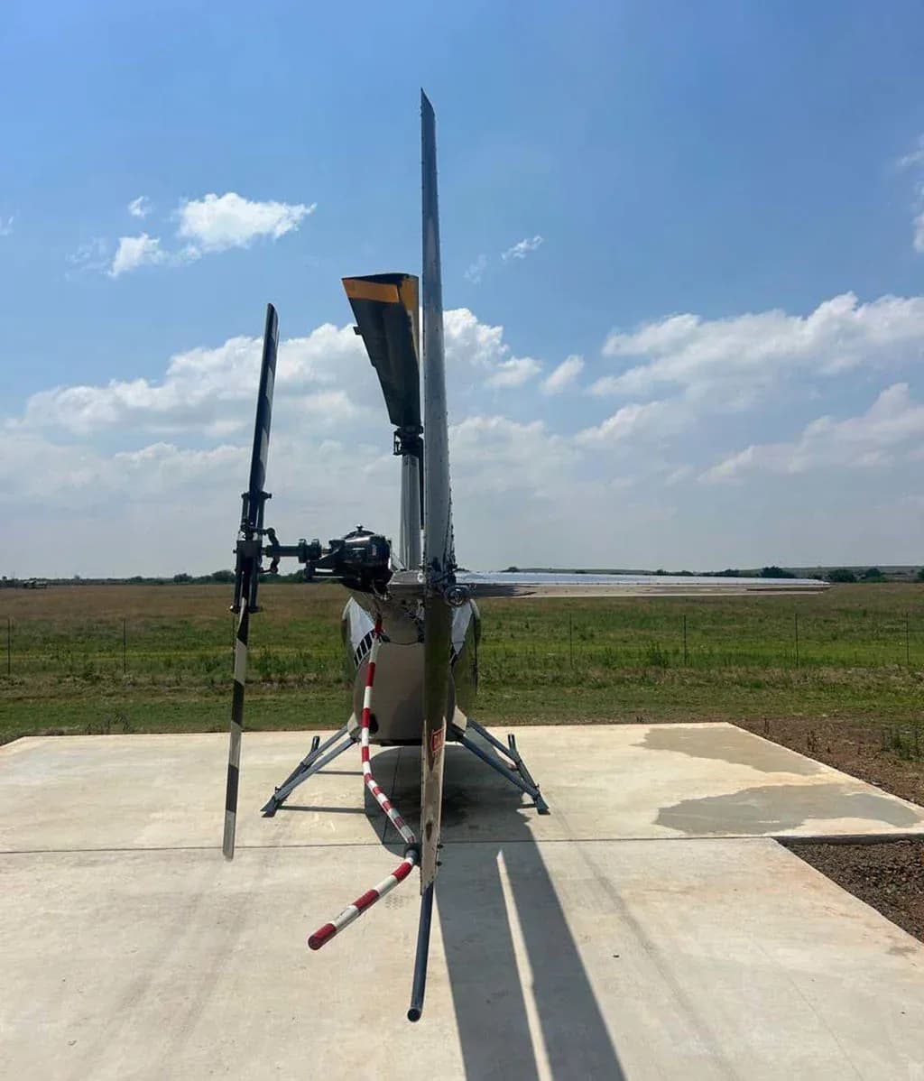 A front view of a silver Robinson R44 helicopter with its rotor blades secured, parked on a concrete pad in a field.
