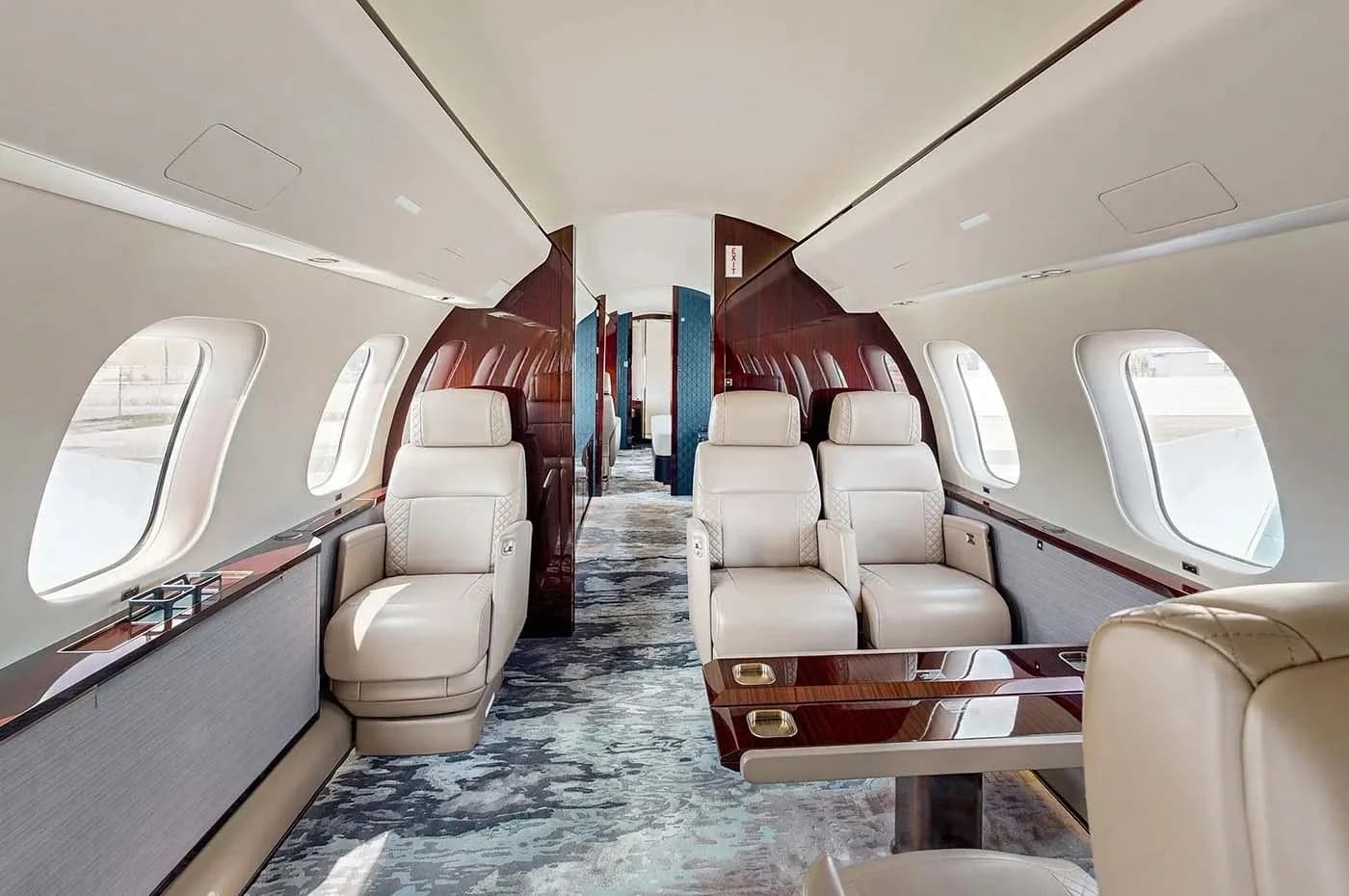 A private jet cabin section with four light beige quilted leather seats facing each other across a polished wooden table, and dark wood accents.