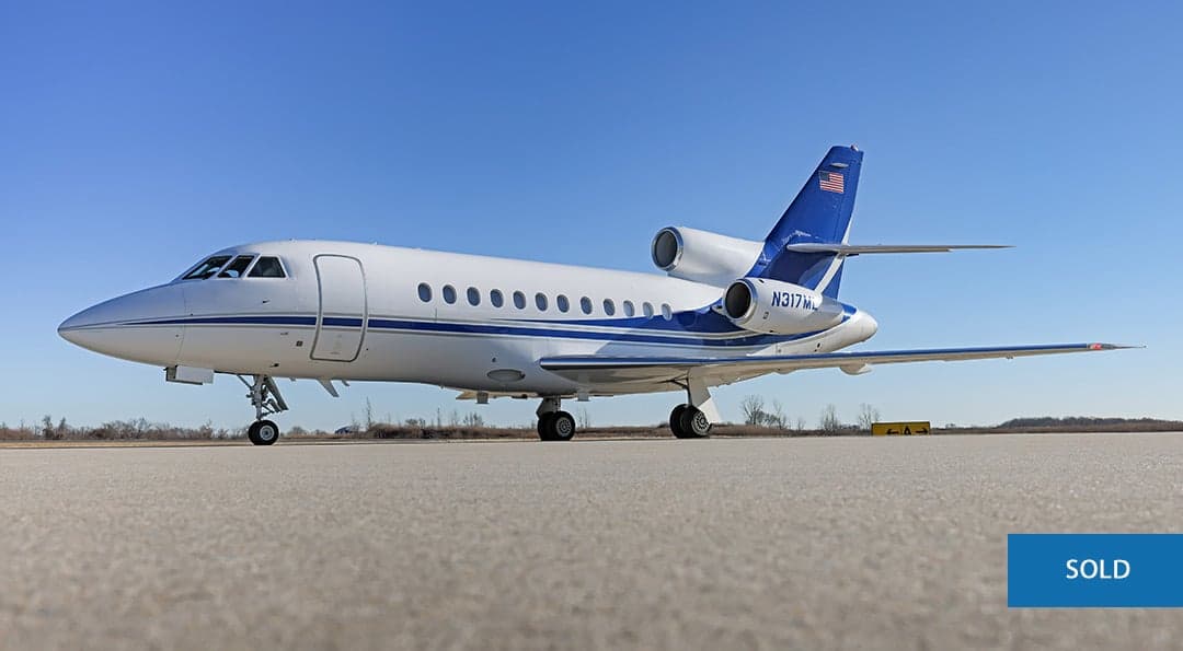 A Dassault Falcon 900EX aircraft is parked on the tarmac with a blue and white paint scheme.