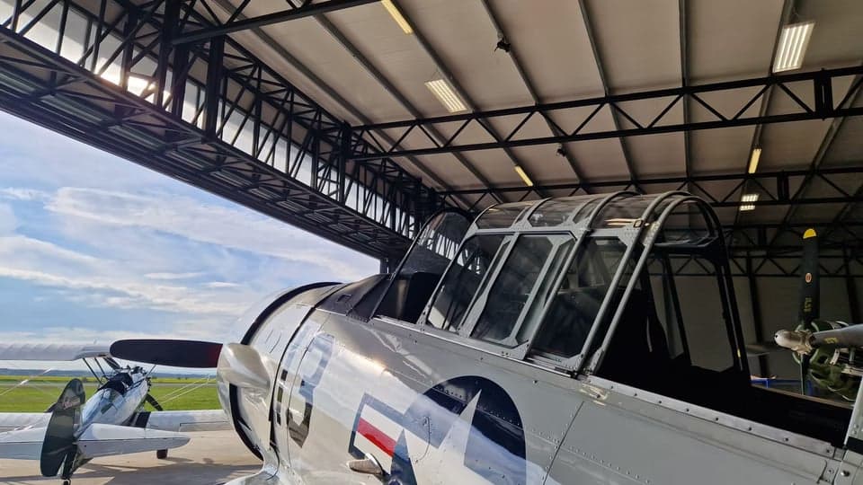 A white and red North American Aviation T-6 Texan aircraft is parked inside a hangar, with the canopy open.