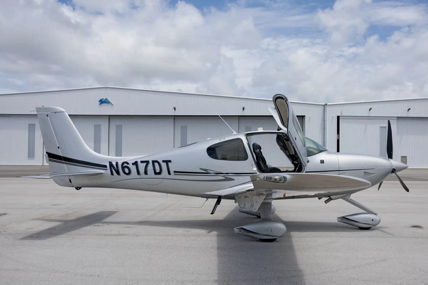 A side view of the silver Cirrus SR22 aircraft with tail number N617DT and its gull-wing doors open, parked in front of hangars.