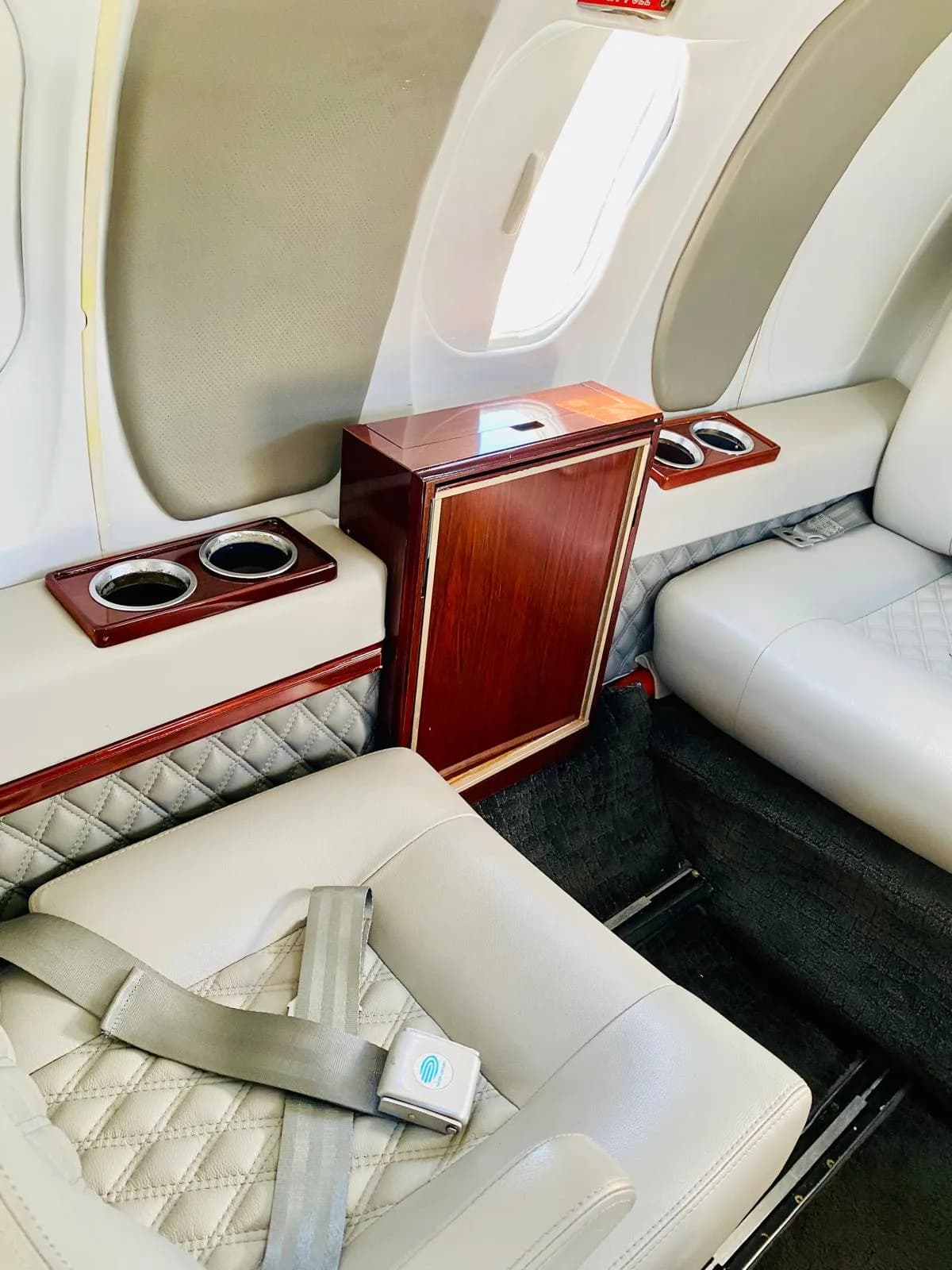 A close-up of a light gray quilted leather seat, side table with cup holders, and a window in the aircraft cabin.