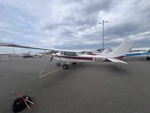 A white and maroon Cessna 182 aircraft (N193R) is parked on an asphalt tarmac with other planes in the background.