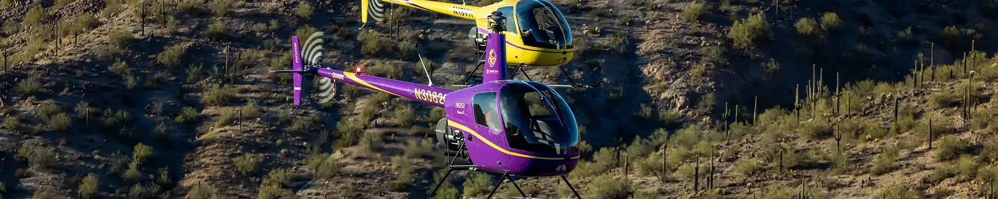 Two Robinson R44 helicopters, one purple and one yellow, flying over a desert landscape.