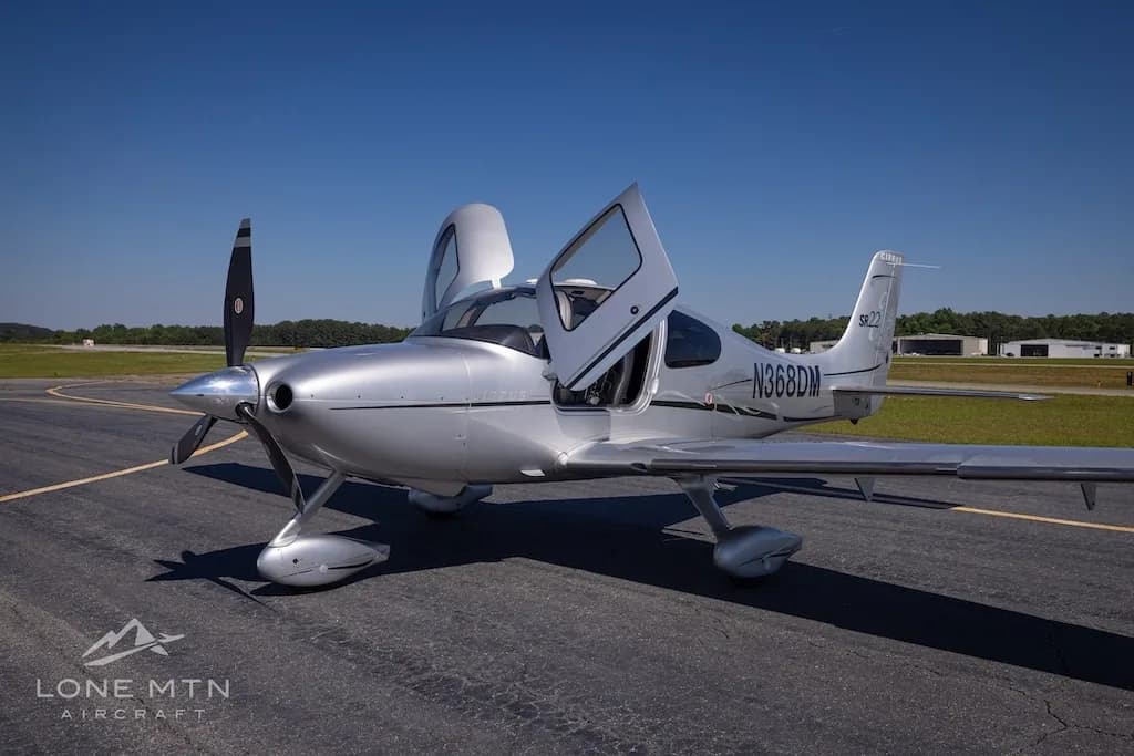 A silver Cirrus SR22 Turbo aircraft (N368DM) with its doors open, parked on an asphalt runway under a clear blue sky.