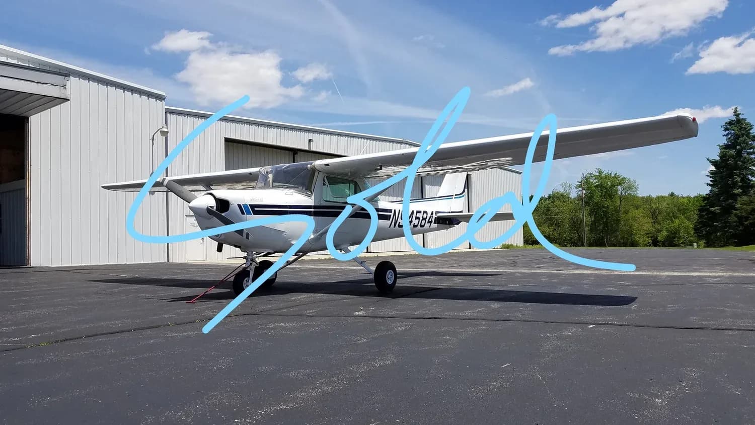 A white Cessna 172 aircraft with blue stripes is parked on an asphalt ramp next to a hangar under a blue sky.