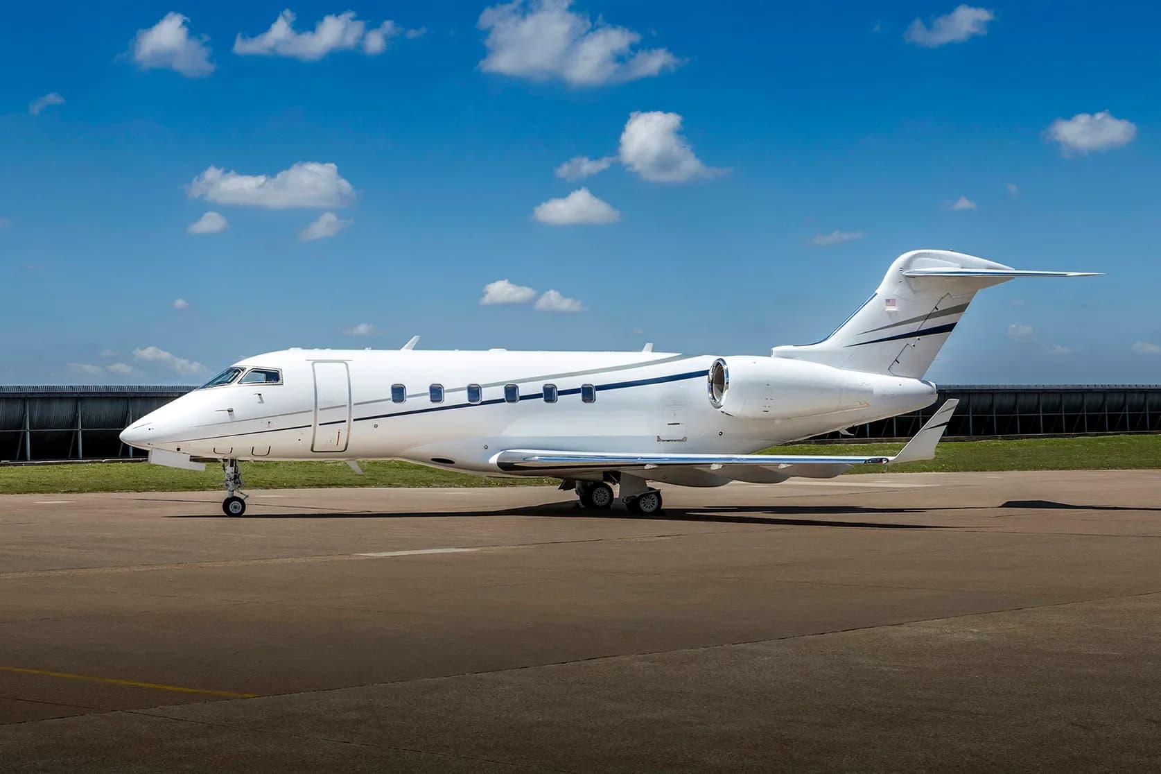 A white business jet with blue and grey stripes is parked on an airfield under a blue sky with scattered clouds.