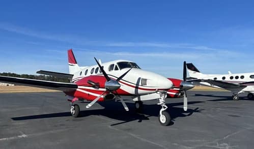 A white and red twin-engine turboprop aircraft, likely a King Air, parked on an asphalt ramp under a clear blue sky, with another aircraft in the background.