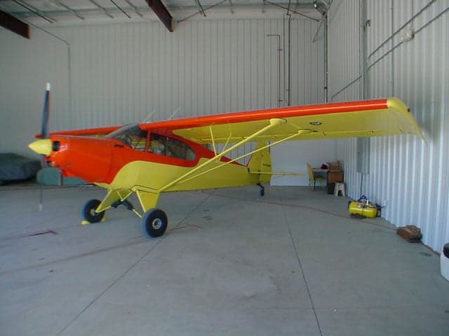 A full side view of an orange and yellow Piper Super Cub aircraft inside a hangar.