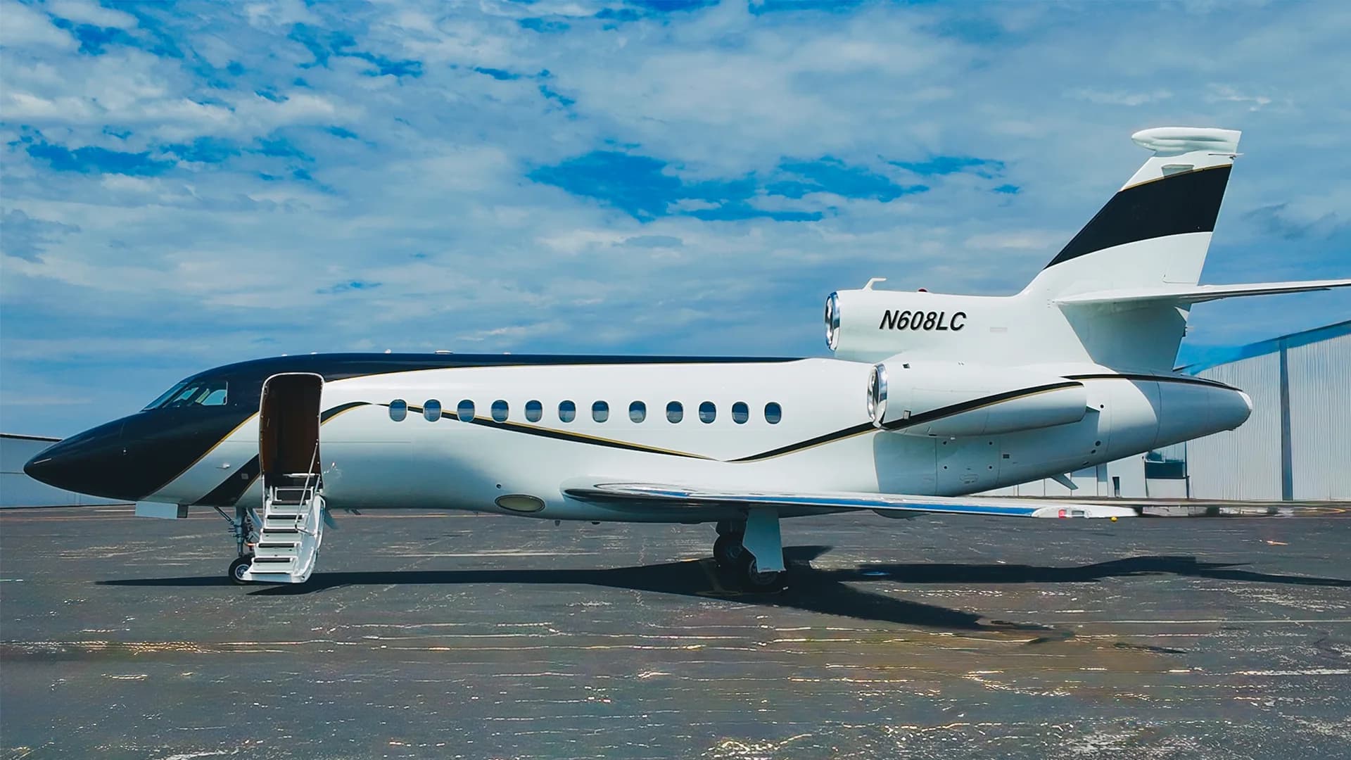 A white and black Dassault Falcon 50 business jet with its door open and stairs extended, parked on an asphalt tarmac under a partly cloudy sky.