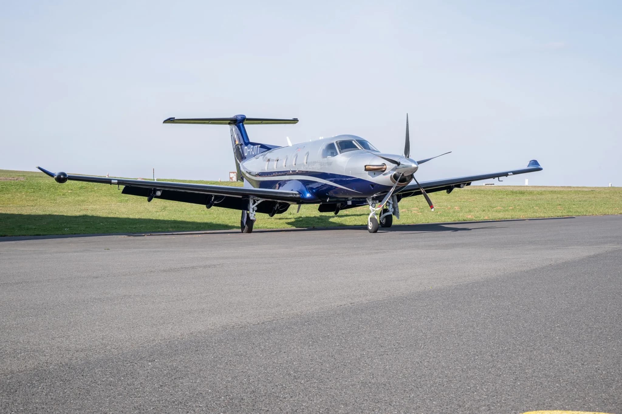 A silver and blue Pilatus PC-12 NG aircraft is parked on an asphalt runway under a clear sky.
