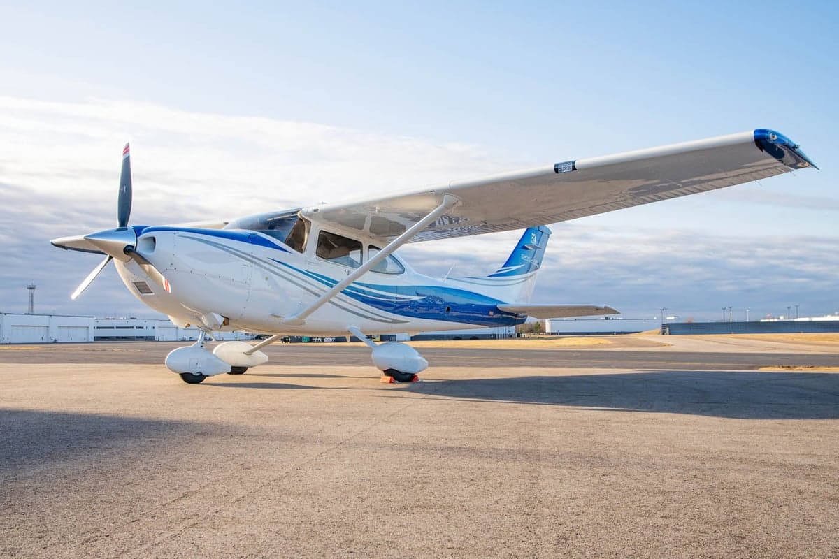 A white and blue Cessna 182 Skylane aircraft parked on an airport tarmac under a partly cloudy sky.
