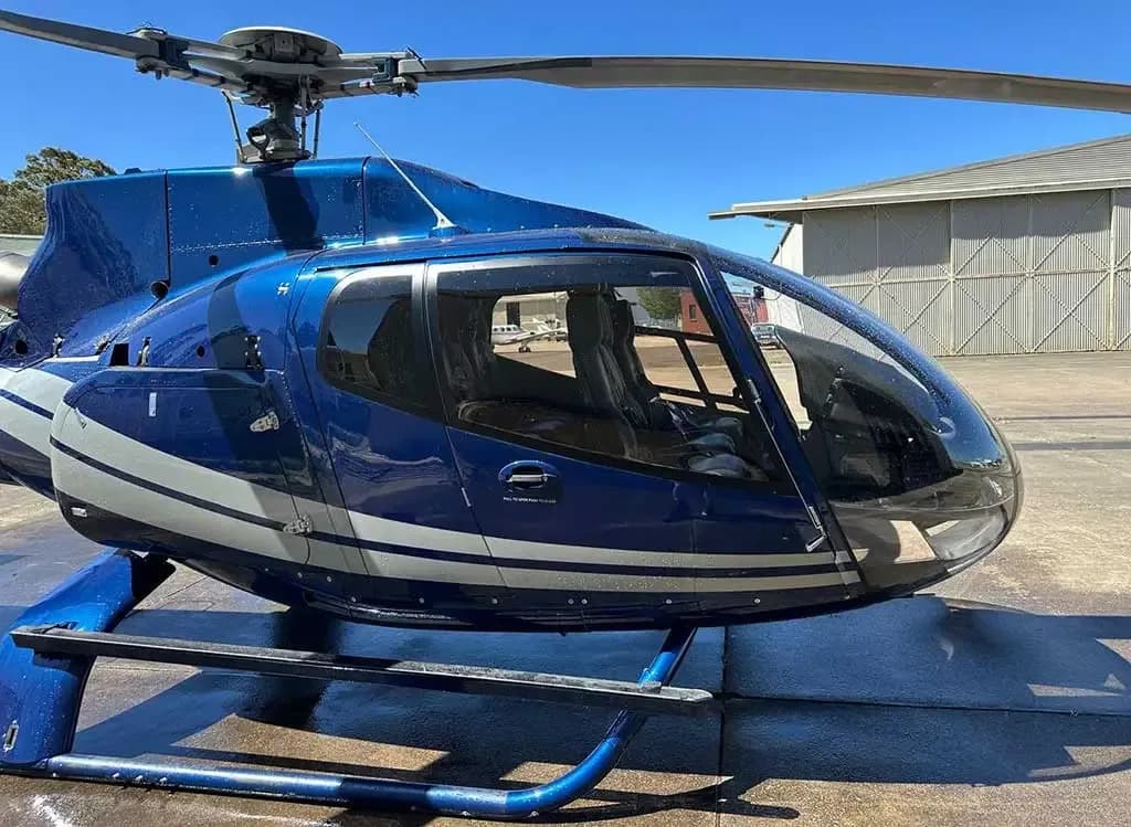 A blue helicopter with white stripes parked on an asphalt tarmac with a hangar in the background.