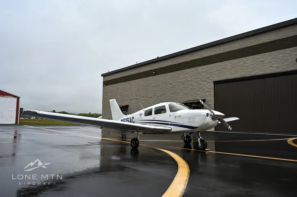 A white single-engine aircraft, N616AC, with blue stripes is parked on a wet tarmac in front of a hangar.