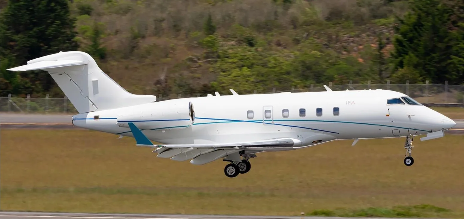 A white business jet with blue and teal stripes, registration "IEA", is captured mid-air during landing or takeoff over a grassy field.