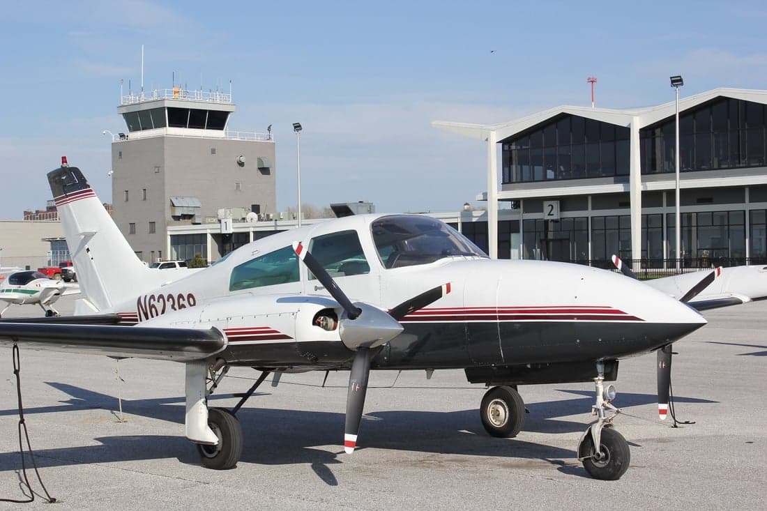 A white and dark gray Piper aircraft is parked on the tarmac in front of a building and control tower.