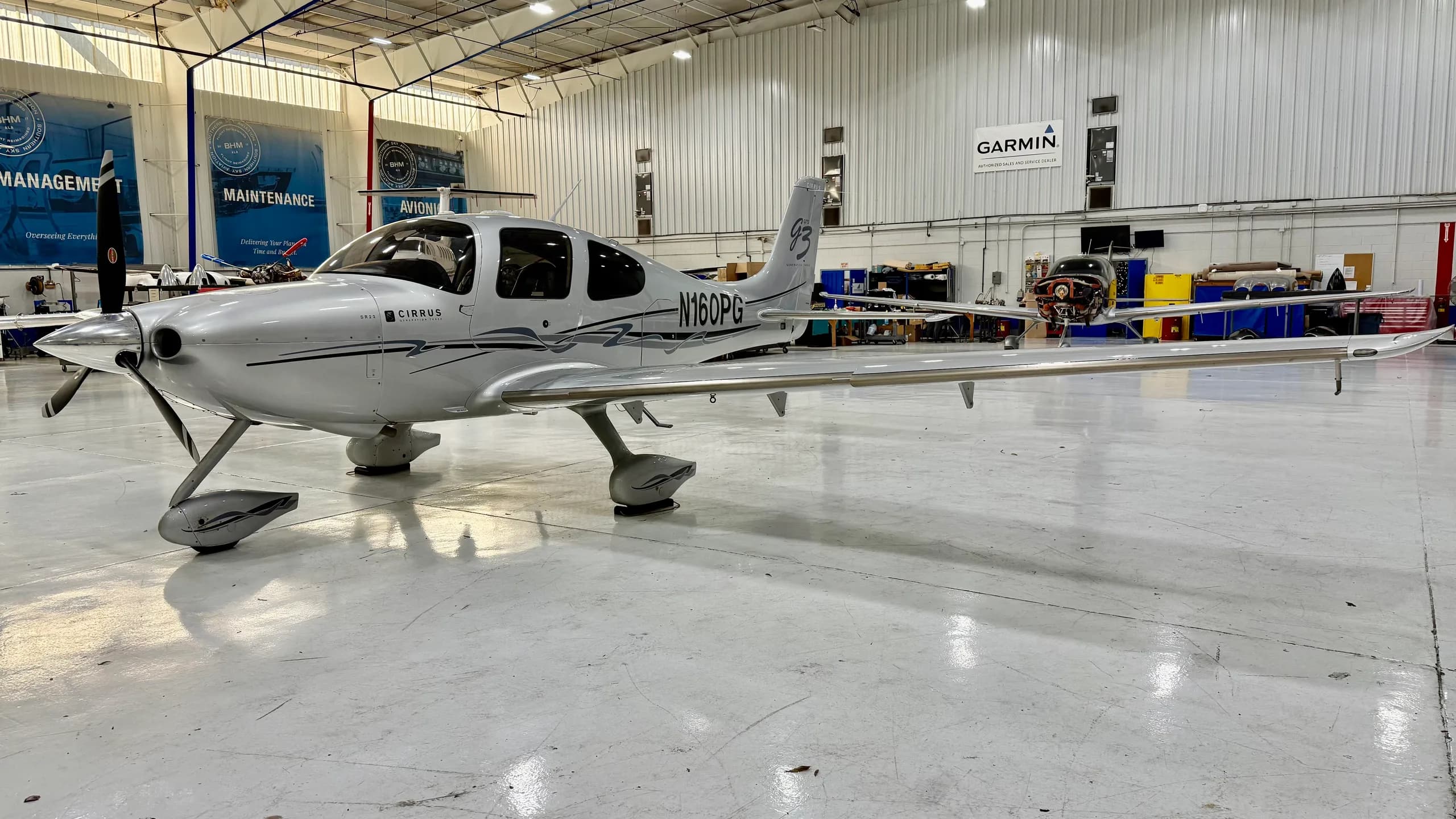 A silver Cirrus SR22 aircraft with dark grey accents, viewed from the front-left in a hangar.