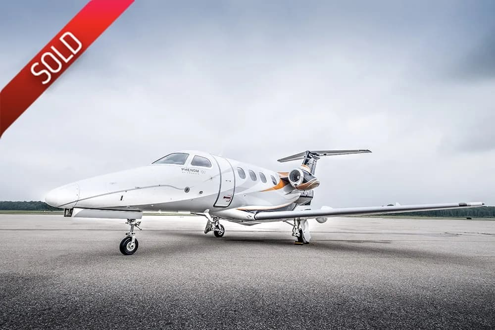 A white Embraer Phenom 300 business jet with orange and grey accents is parked on an airport tarmac under a cloudy sky.