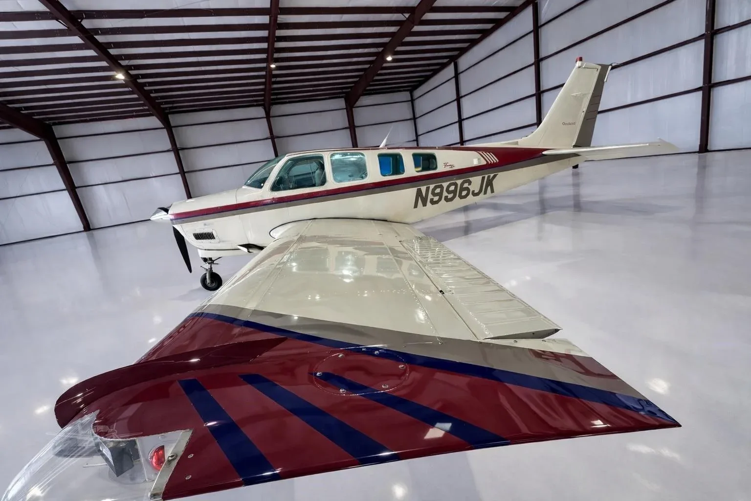A white Beechcraft Bonanza with red and blue stripes on the wings and tail, parked in a hangar.