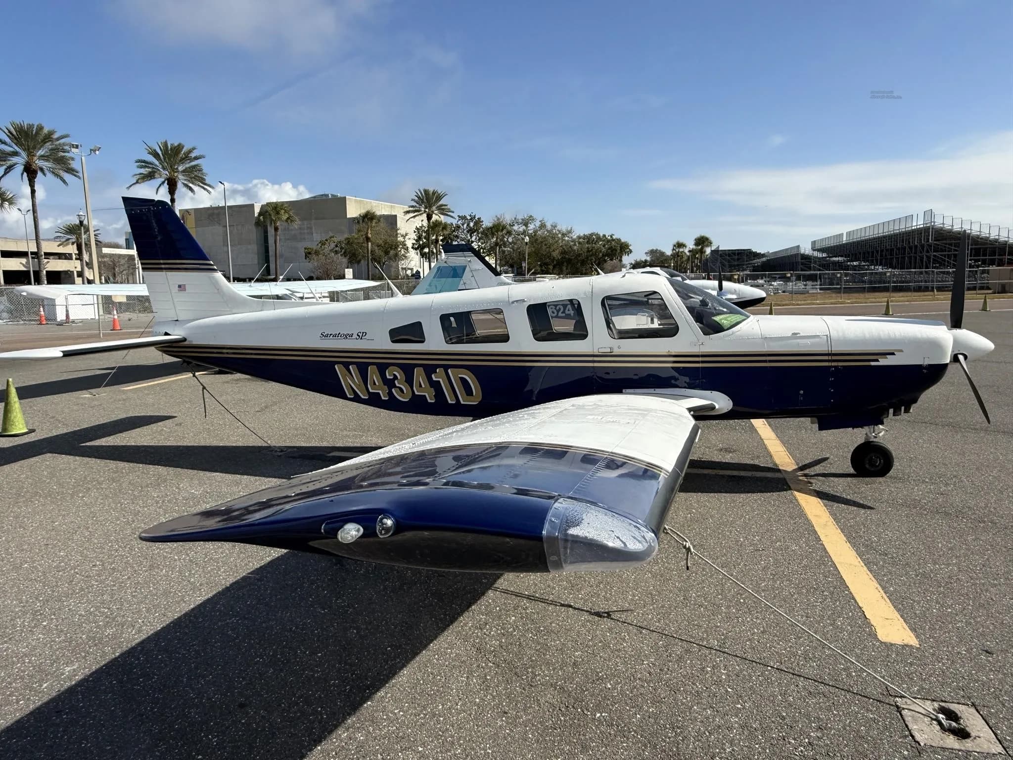 A blue and white Piper Saratoga SP aircraft (N4341D) parked on an asphalt tarmac with palm trees in the background.