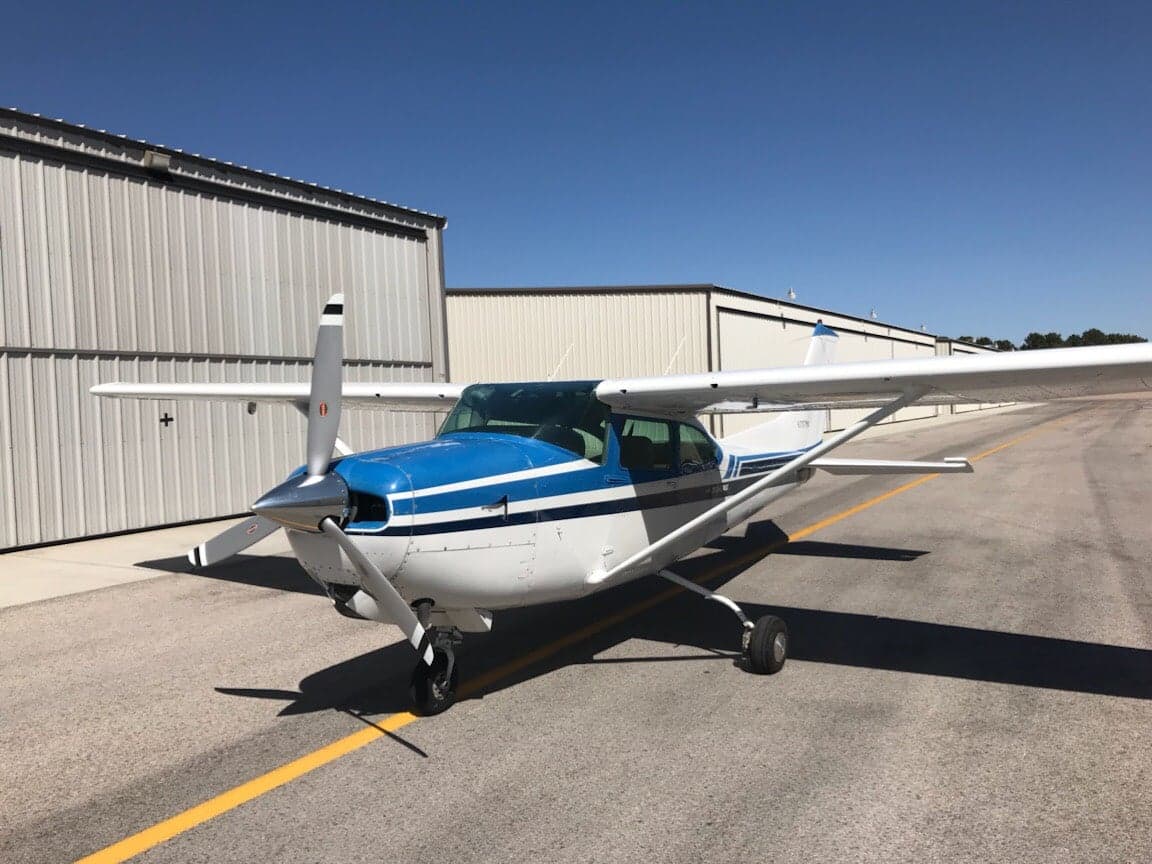A white and blue single-engine propeller aircraft is parked on an asphalt ramp in front of hangars under a clear sky.
