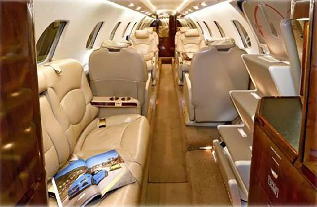 The luxurious interior cabin of a private jet, featuring light beige leather seats and dark wood accents.