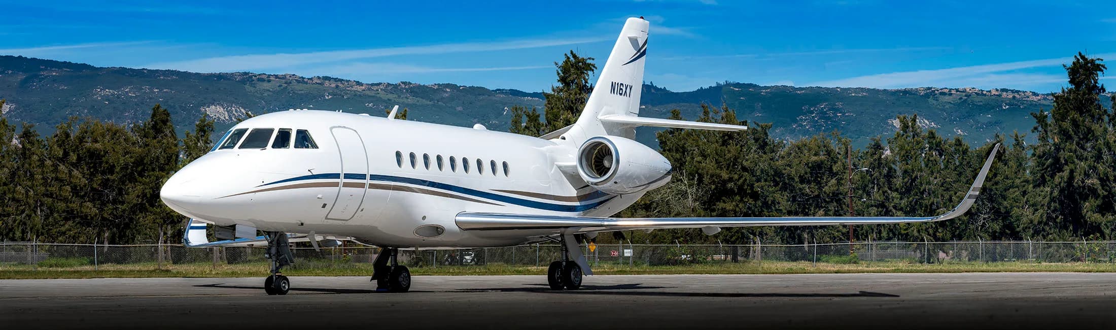 A white Falcon aircraft with blue and gold stripes sits on the tarmac with trees and mountains in the background.