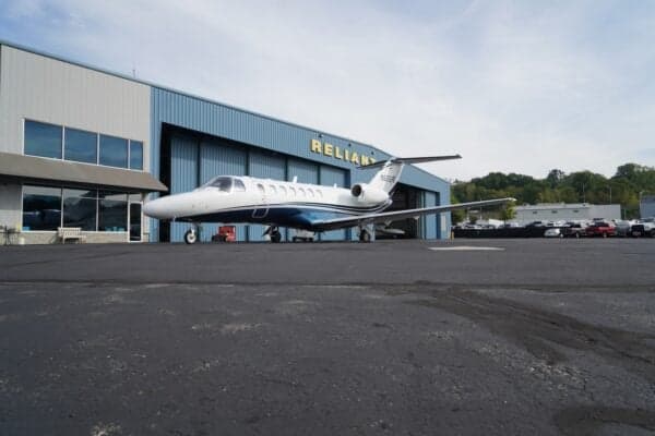 A white business jet with a blue stripe is parked on an asphalt apron in front of a blue hangar labeled 'RELIANT'.