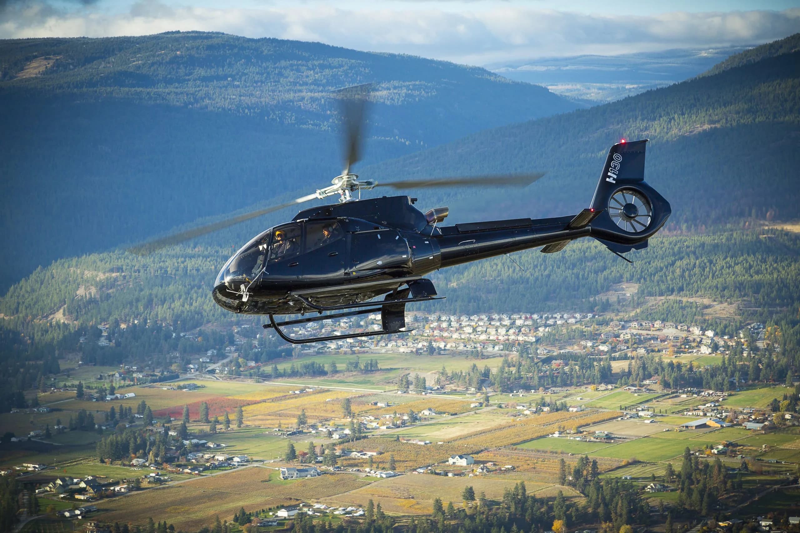 A black Airbus H130 helicopter with two pilots flying over a scenic valley with vineyards, houses, and forested mountains.