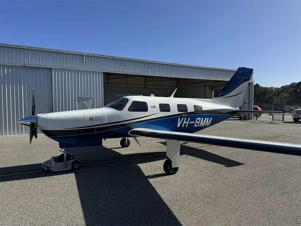 A white and blue Piper M350 aircraft, registration VH-8MM, is parked on an asphalt tarmac next to a hangar under a clear blue sky.