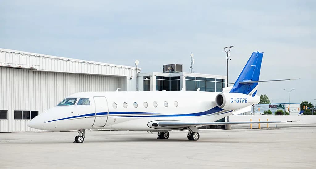 A white business jet with blue stripes and tail, registration C-GTRG, parked on an airport tarmac next to a hangar.