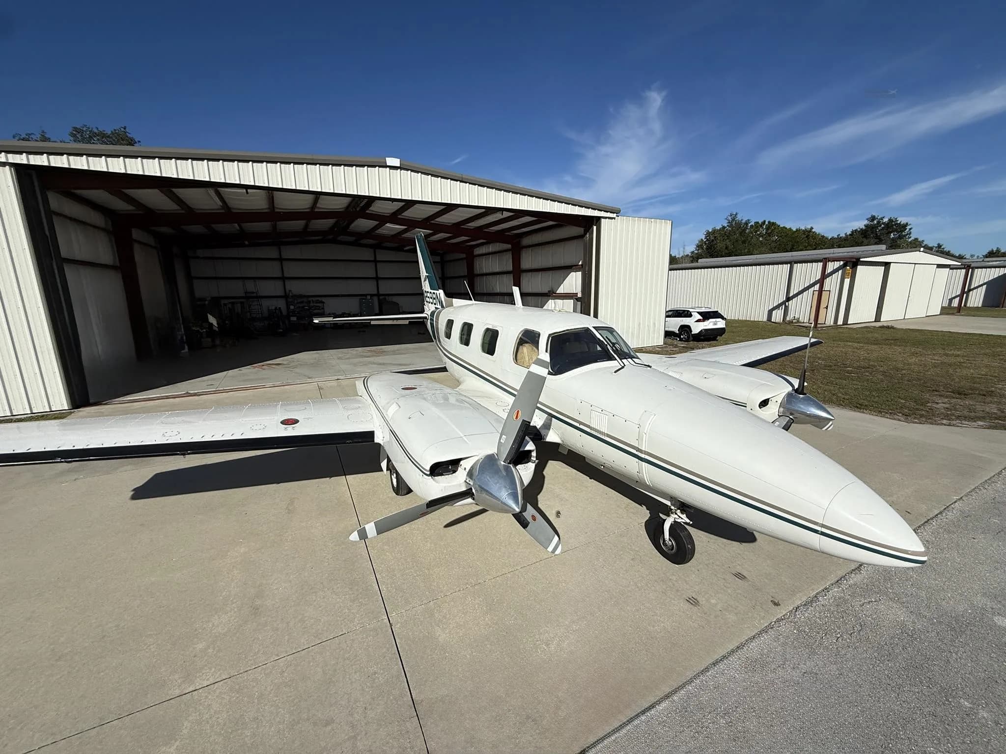 A white twin-engine propeller aircraft with a green stripe is parked on a tarmac in front of an open hangar under a blue sky.