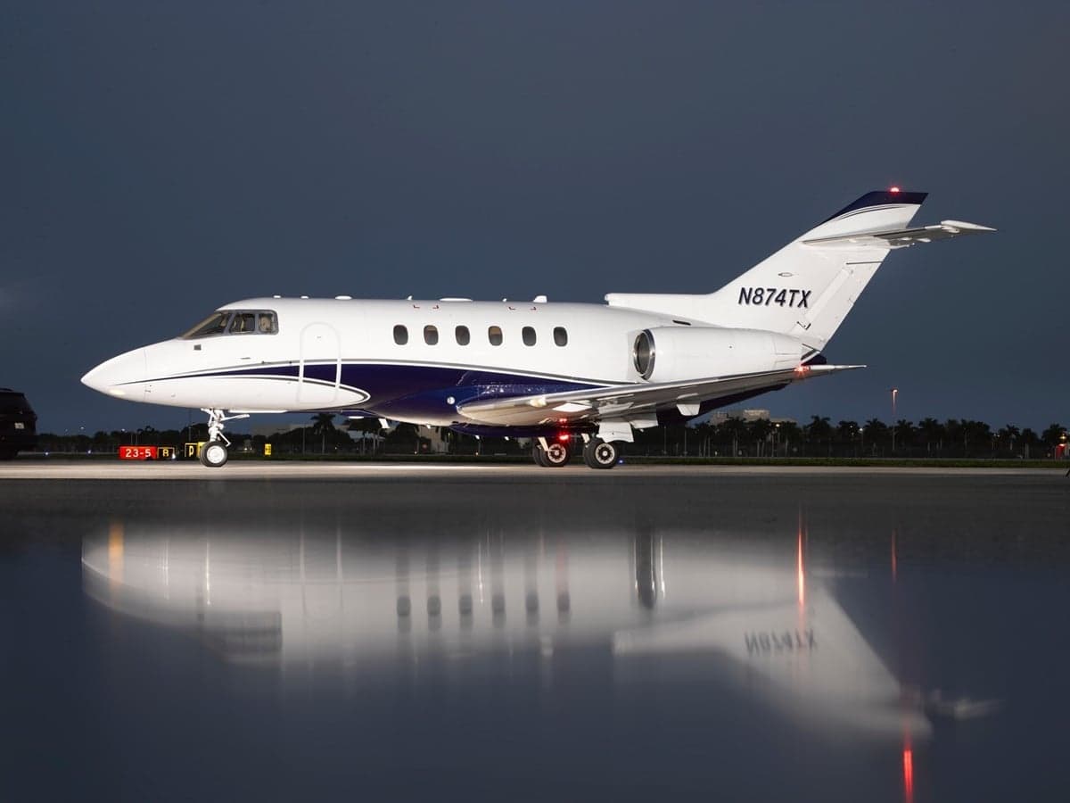 A white and blue business jet (N874TX) is parked on a wet tarmac at night, reflecting its image.